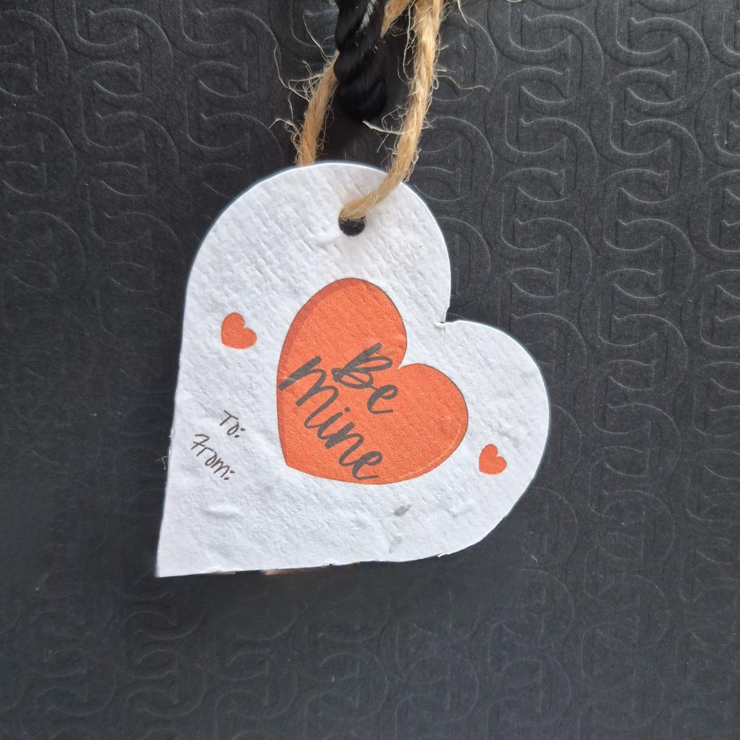 Valentine's day  heart-shaped plantable seed paper gift tag with 'Be mine' text. Hanging from a  black bag
