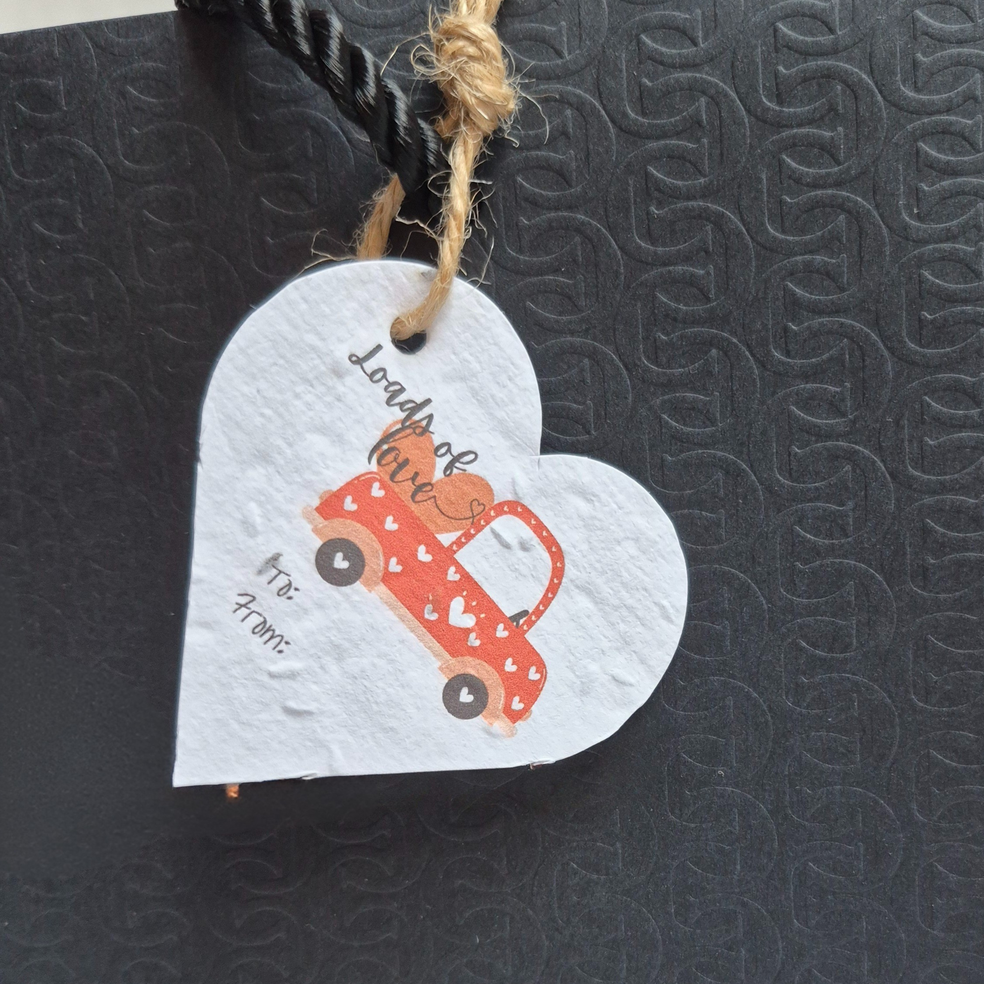  Valentines day heart-shaped seed paper gift tag with a red truck with a truckload filled with 'loads of love' text. Hanging from a black bag.