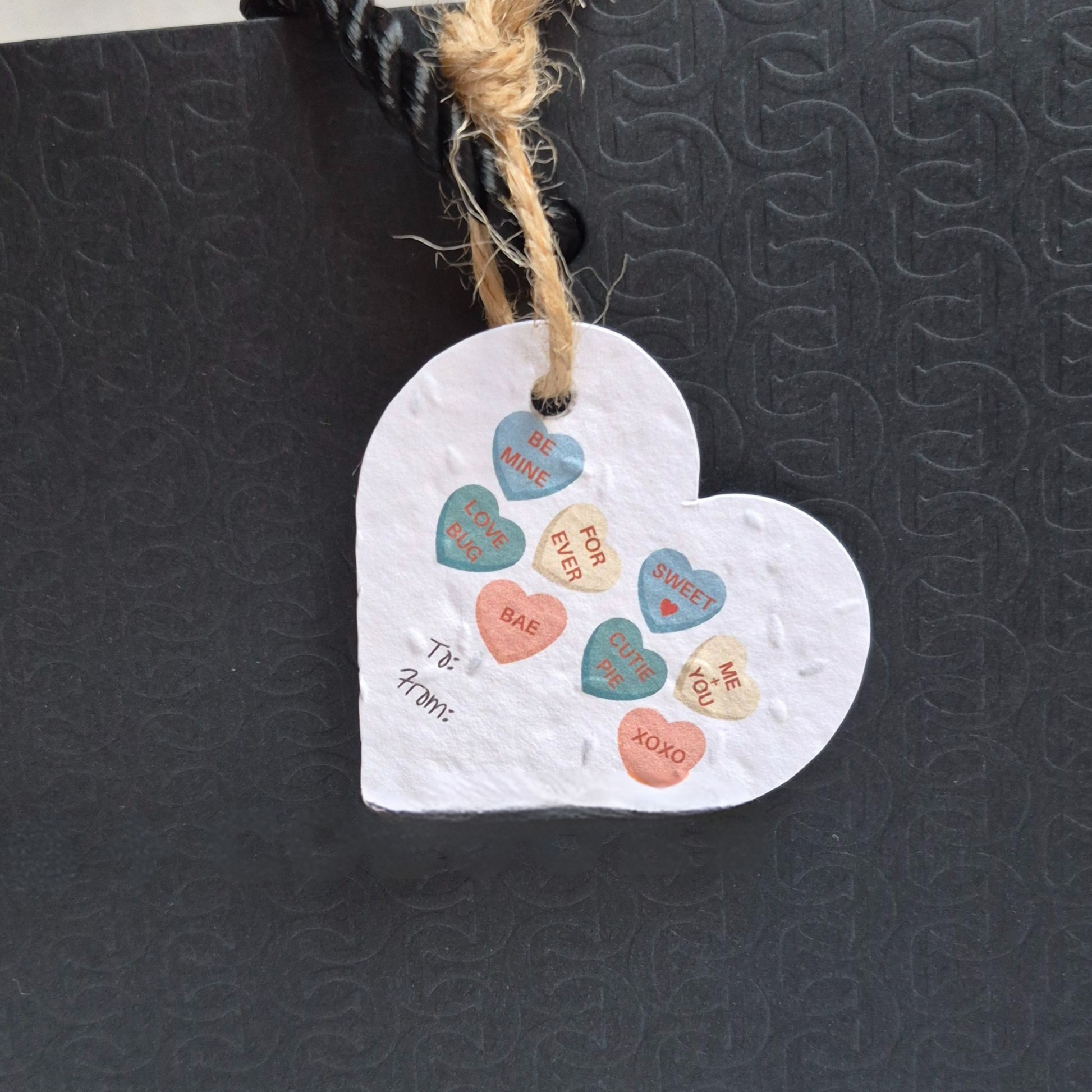Valentines day heart-shaped seed paper gift tag with candy heart designs. Hanging from a black bag.