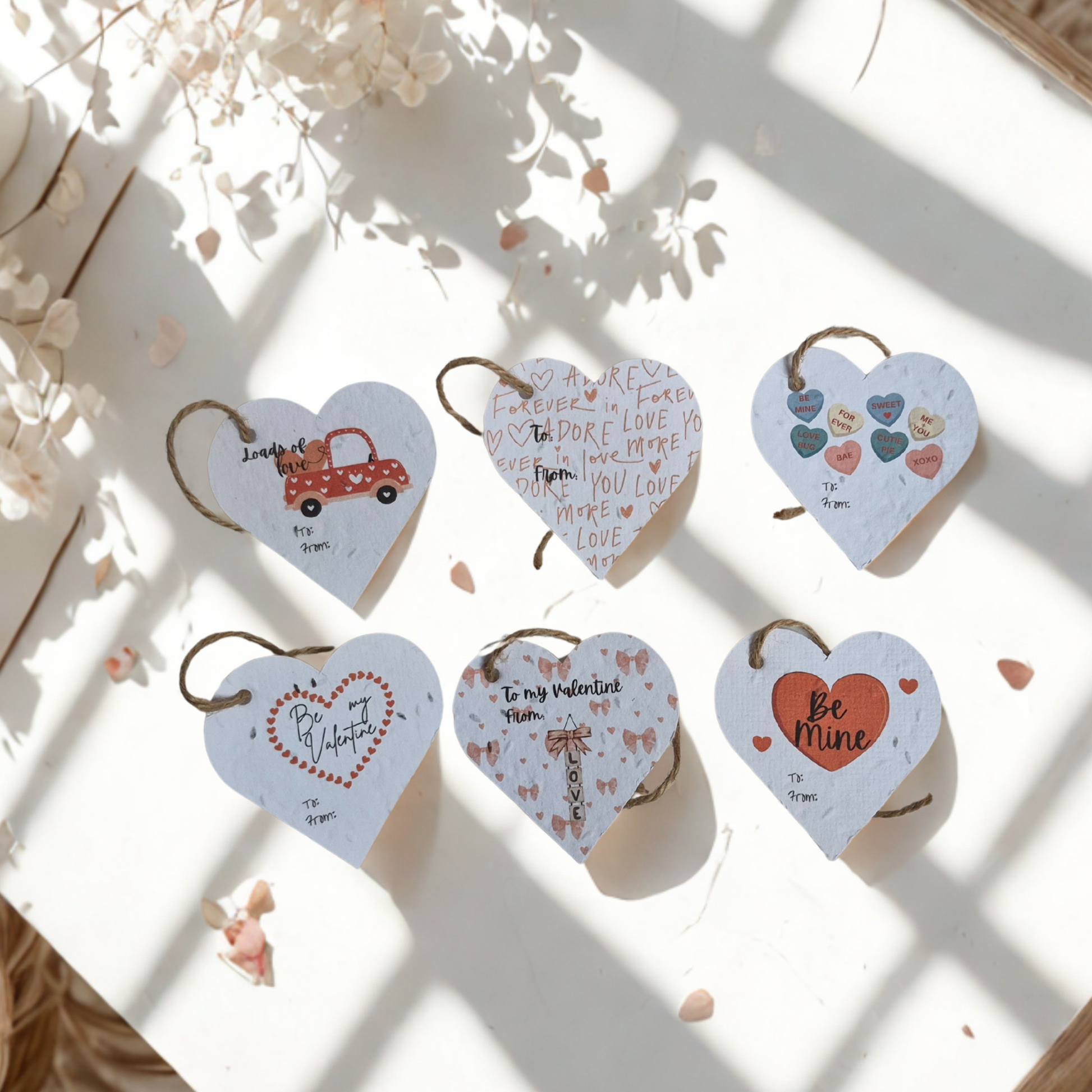 Set of six Heart-shaped gift tags with various Valentines day messages, mad eof seed paper, placed on a light surface with flowers.