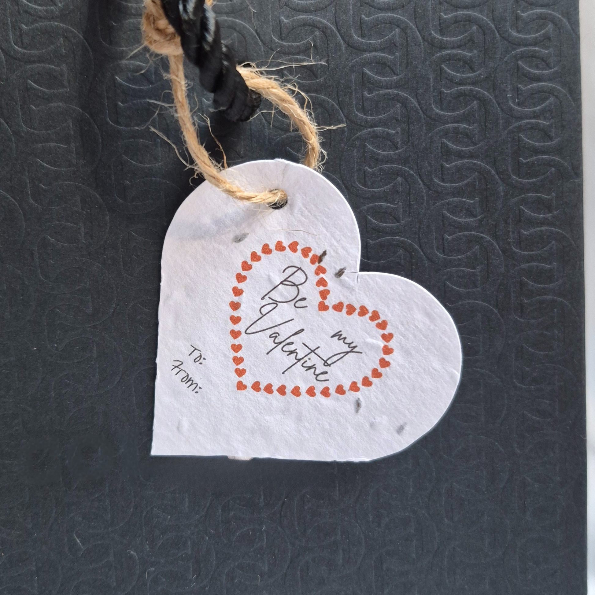 Valentines day heart-shaped seed paper gift tag with 'Be my Valentine' text. Hanging from a black bag.